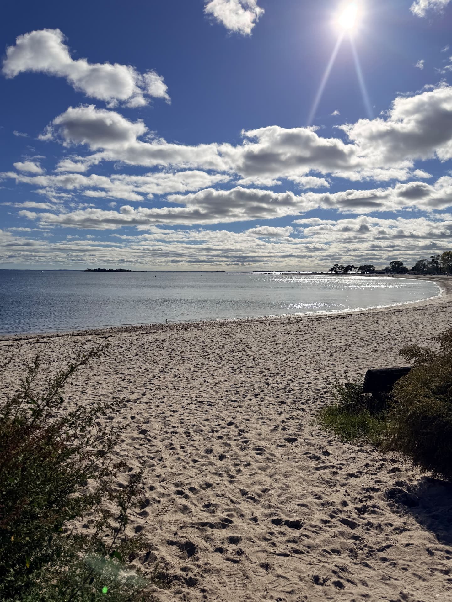 Sandy beach curving around calm, sunlit water under scattered clouds