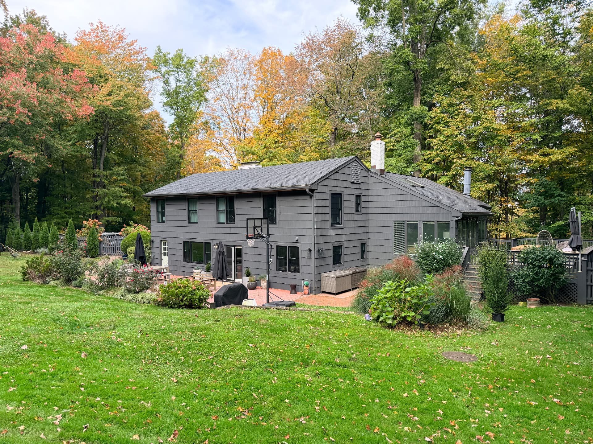The back of the house after the work: charcoal siding and dark windows among autumn trees, with the patio and lawn