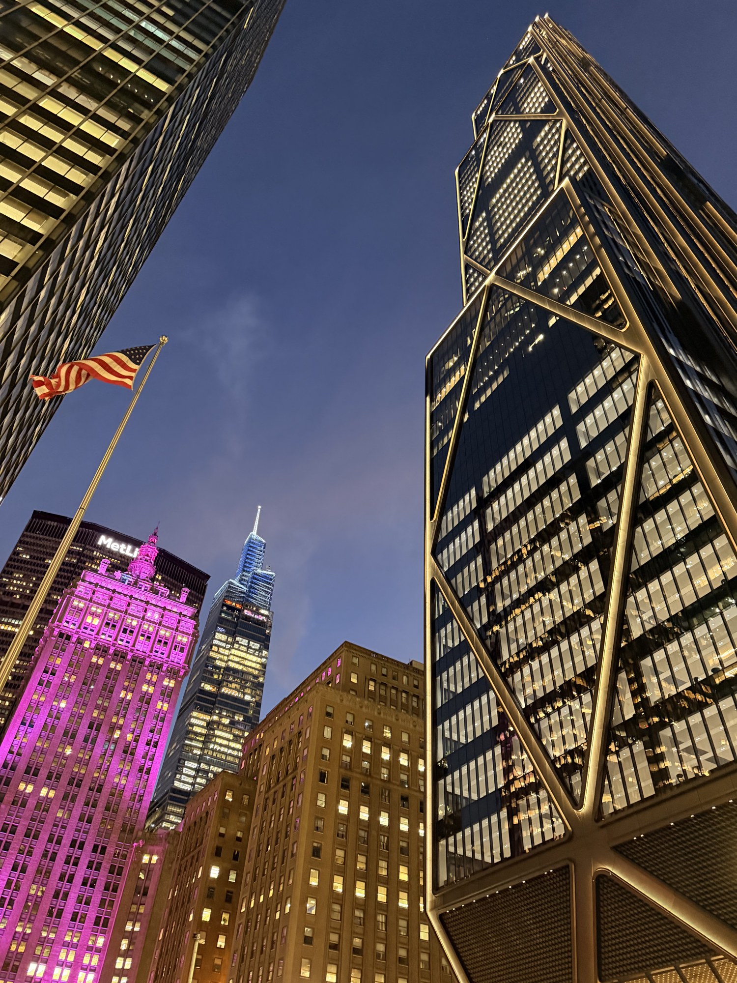 Looking up at lit Midtown towers and an American flag against a dusk sky
