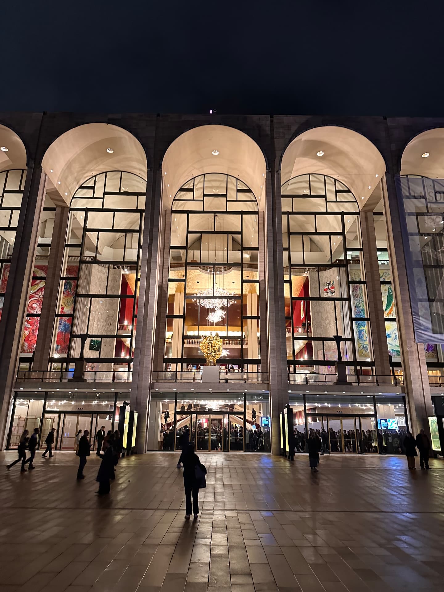 The Metropolitan Opera House at Lincoln Center lit at night, its tall arched windows showing chandeliers and murals, people on the plaza