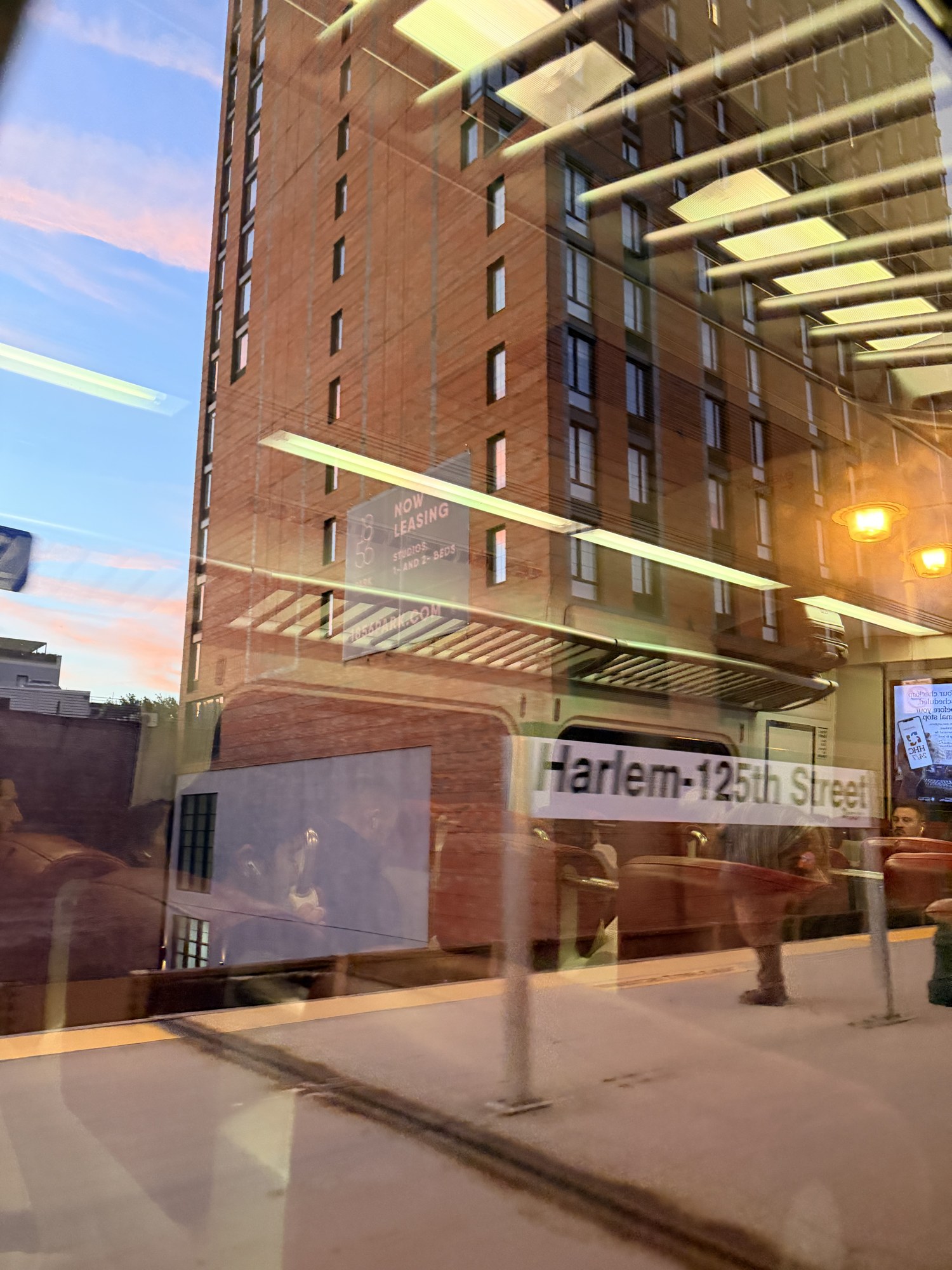 Harlem–125th Street station sign and a brick tower reflected in a train window at sunset