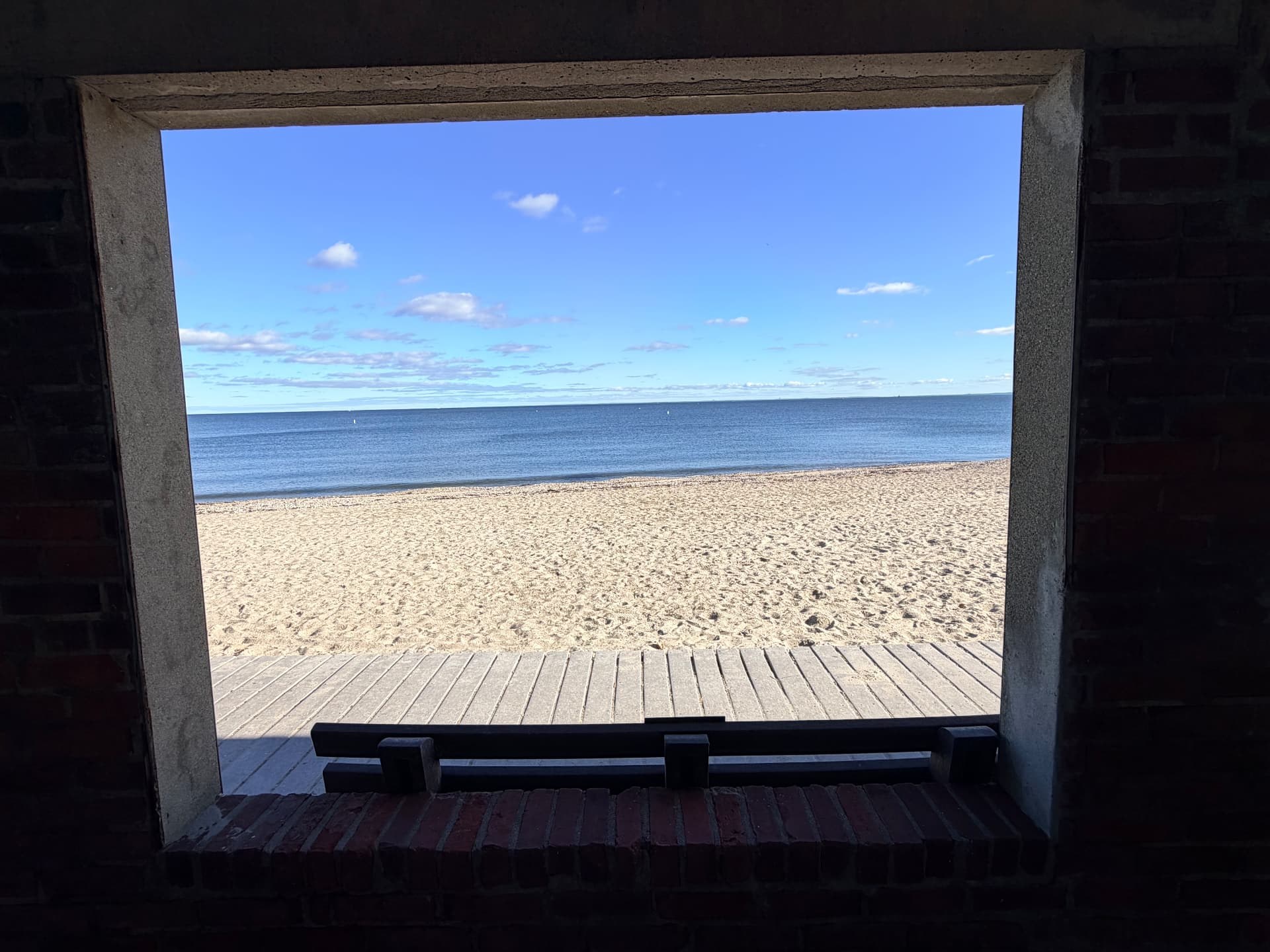 Looking through an open brick window frame onto a boardwalk, sand and calm water under a blue sky