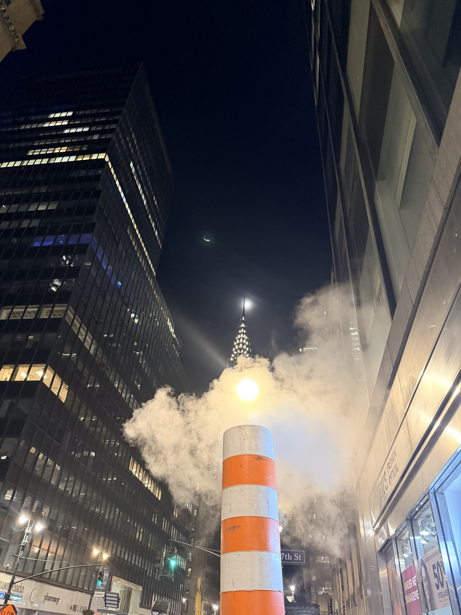 Steam rising from an orange and white street stack, with the Chrysler Building spire behind at night