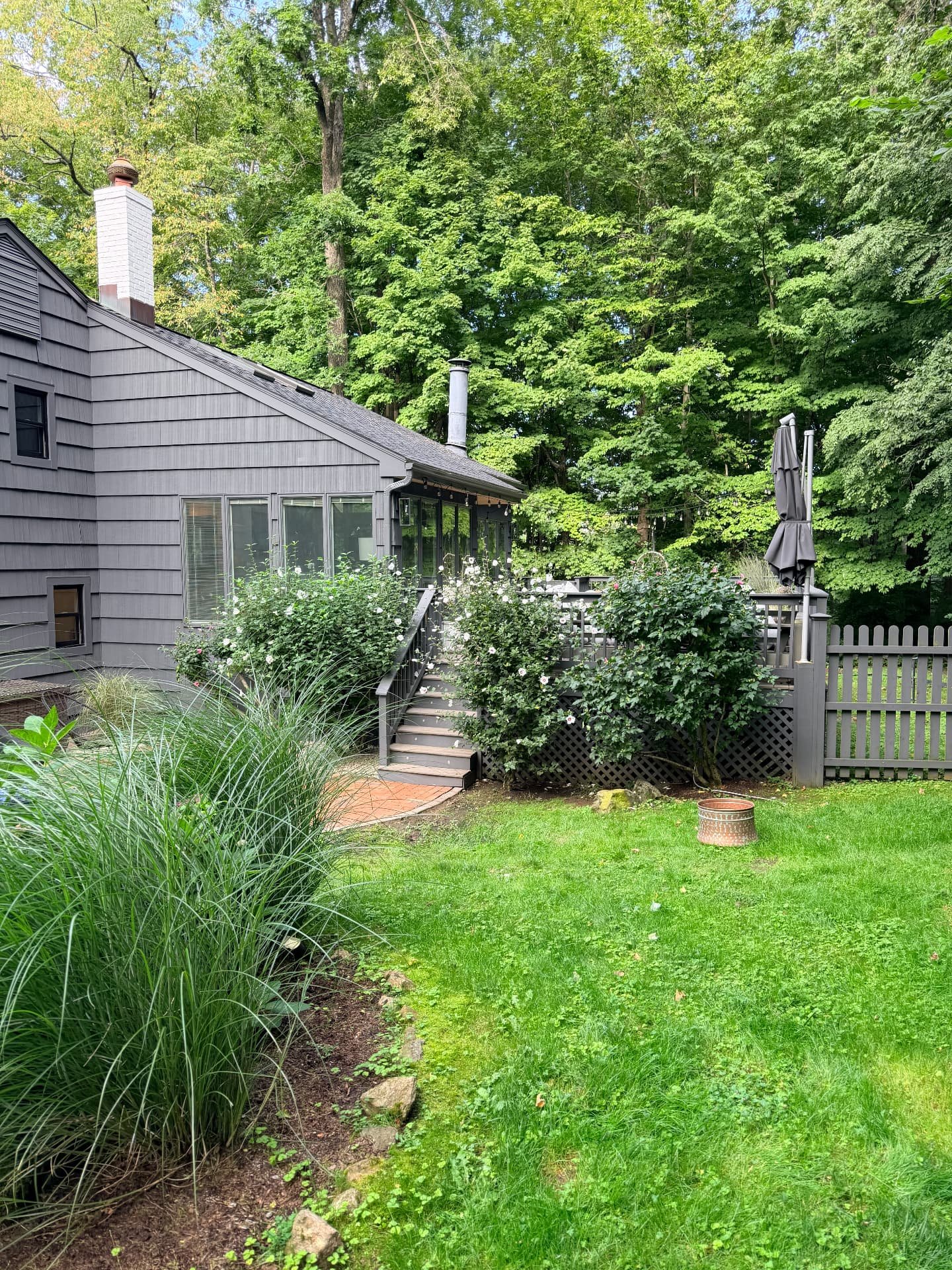Charcoal-sided house beside a deck with flowering shrubs, grasses and tall green trees